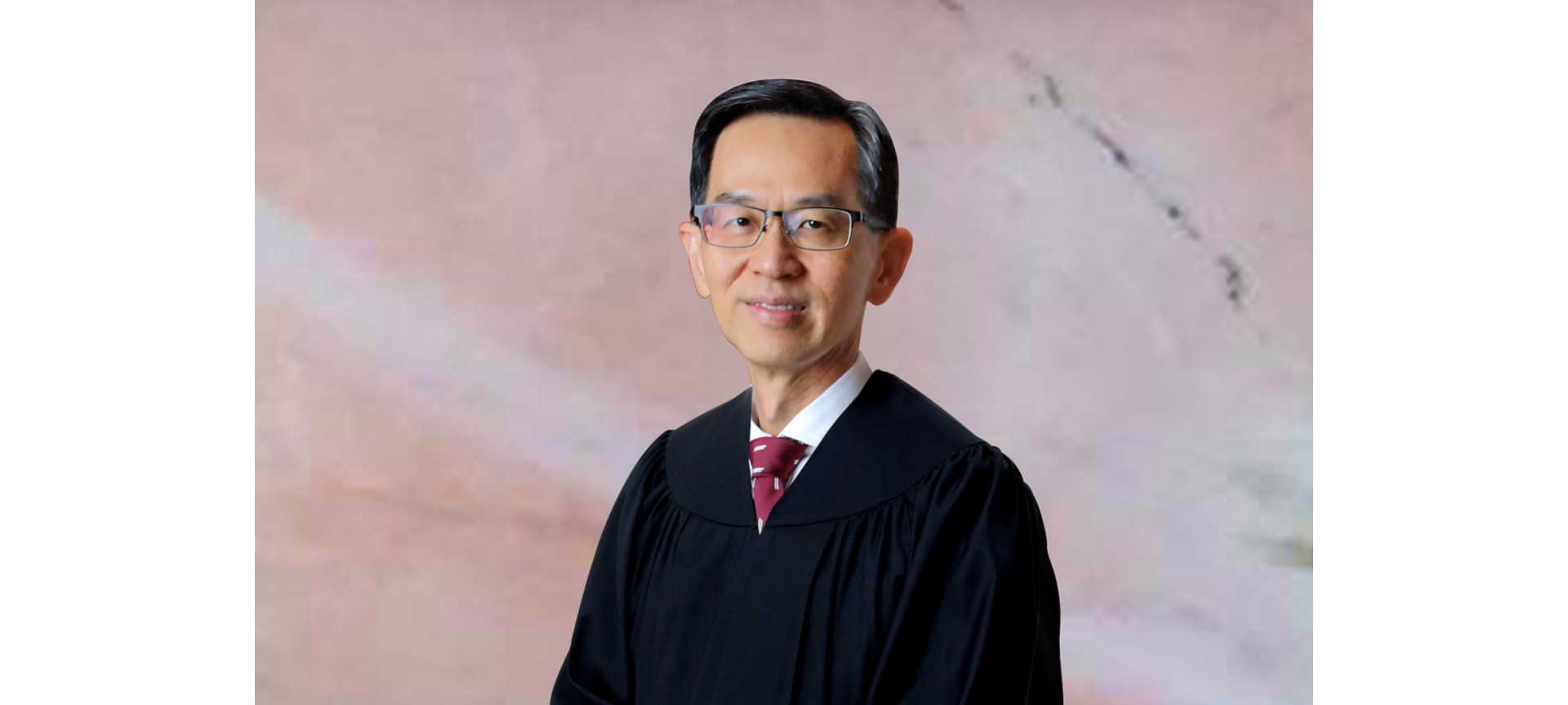 A man wearing a black judicial robe, white dress shirt, and red tie, against a pink marbled background.